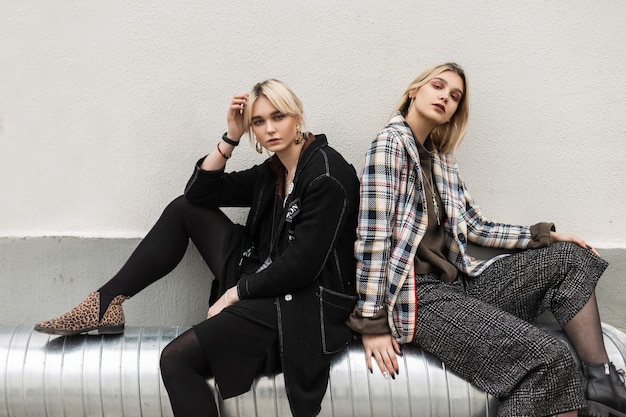 Twee mooie jonge zussen met blond haar met sexy lippen in vintage kleding in mode-laarzen ontspant op metalen pijp in de buurt van de muur in de stad