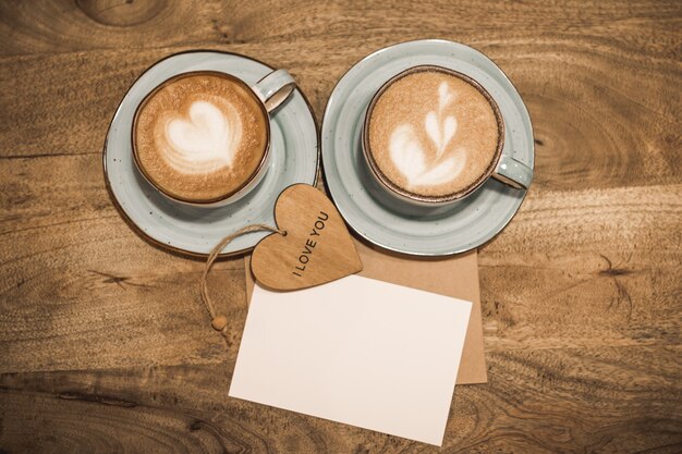 Twee mooie kopjes koffie met een hartvorm, ambachtelijke envelop, wit blanco vel papier en houten hart met de woorden ik hou van jou op een houten achtergrond. Valentijnsdag concept. Selectieve aandacht.