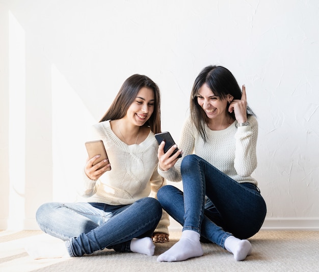 Twee mooie lachende vrouwen in spijkerbroek en witte truien texting naar vrienden met behulp van mobiele telefoon