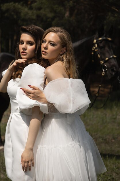 Foto twee mooie meisjes in trouwjurken staan naast een paard in het bos