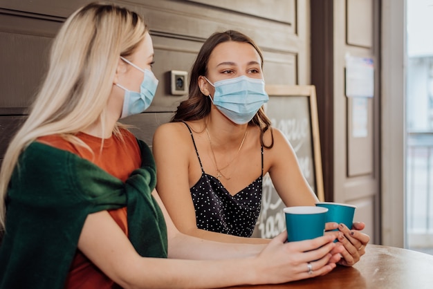 Twee mooie meisjes met maskers op hun gezicht brengen samen tijd door in koffie en thee te drinken