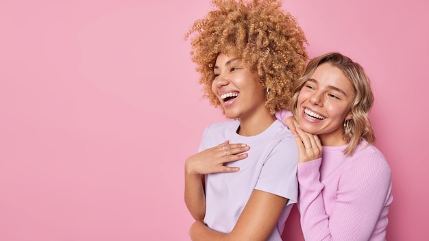 Twee mooie positieve vrouwen lachen vrolijk kijken ergens opzij hebben een vrolijke stemming gekleed in vrijetijdskleding geïsoleerd over roze achtergrond kopie ruimte voor uw reclame-inhoud gelukkig emoties concept