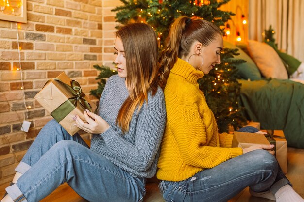 Twee mooie vrolijke gelukkige jonge meisjes vriendin geven kerstcadeaus op de achtergrond van een nieuwe jaarboom thuis