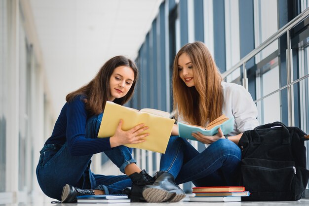 Twee mooie vrouwelijke studenten die met boeken op de vloer in de universitaire gang zitten