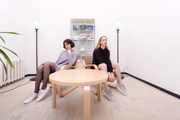 Twee mooie vrouwelijke studenten zitten bij de tafel in een kamer met een minimalistisch interieur.