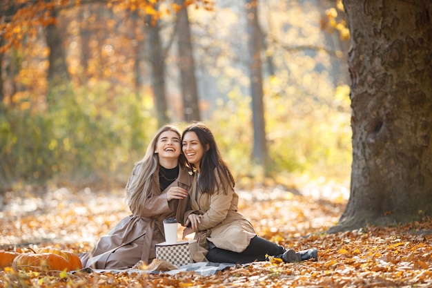 Twee mooie vrouwelijke vrienden die tijd doorbrengen op een picknickdeken op het gras. Twee jonge lachende zussen maken picknick croissants eten in herfst park. Donkerbruine en blonde meisjes die jassen dragen.