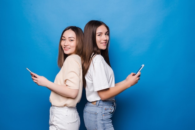 Twee mooie vrouwen die celtelefoons over blauwe muur gebruiken
