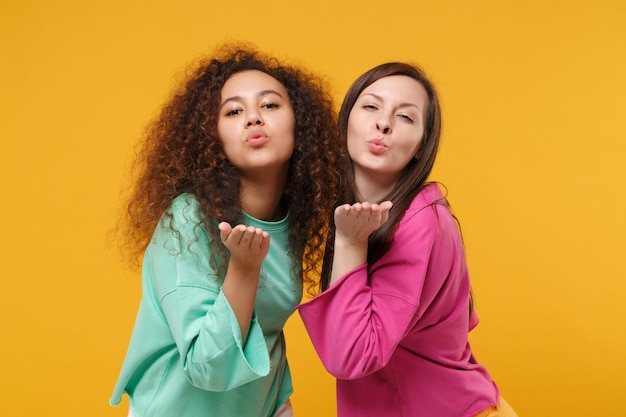 Twee mooie vrouwen vrienden Europese en Afrikaanse Amerikaanse meisjes in roze groene kleding poseren geïsoleerd op geeloranje achtergrond. Mensen levensstijl concept. Bespotten kopie ruimte. Blazen luchtkus verzenden.
