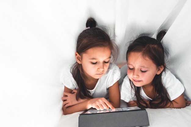 Twee mooie zusjes liggen in bed en kijken naar het scherm van een tablet smart kids