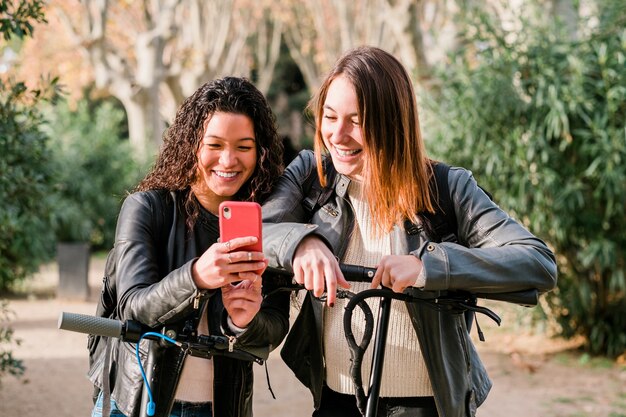 Twee multi-etnische vriendinnen op elektrische scooters die smartphone buitenshuis gebruiken