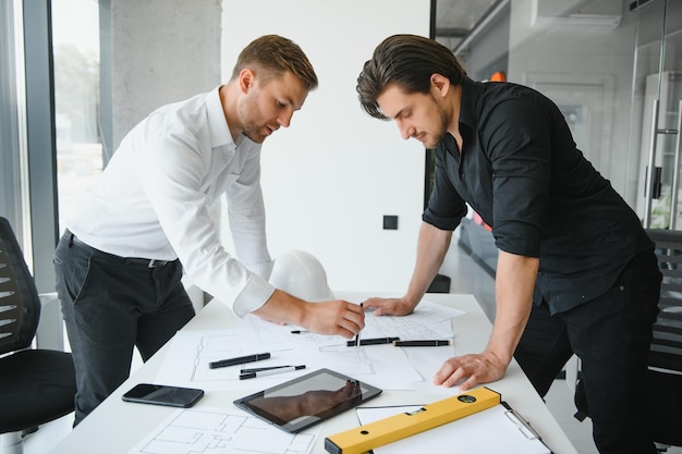 Twee ontwerpers van gebouwen staan in een modern kantoor, leunend over een bureau en bespreken samen blauwdrukken