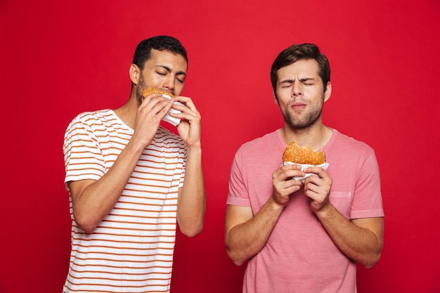 Twee opgetogen mannenvrienden die zich geïsoleerd over rode muur bevinden, hamburgers eten