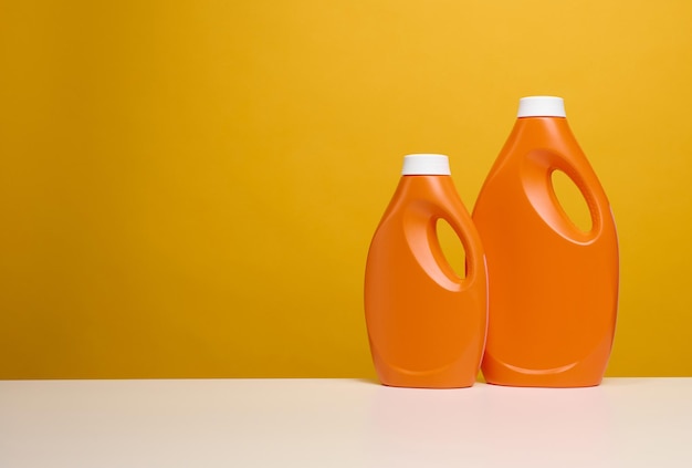 Twee oranje plastic flessen met vloeibaar wasmiddel staan op een witte tafel, gele achtergrond