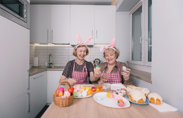 Twee oudere vrouwen met konijnenoren op hun hoofd schilderen thuis in de keuken paaseieren