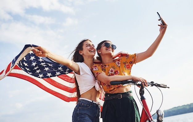 Twee patriottische vrolijke vrouwen met fiets en Usa-vlag in handen maken selfie.