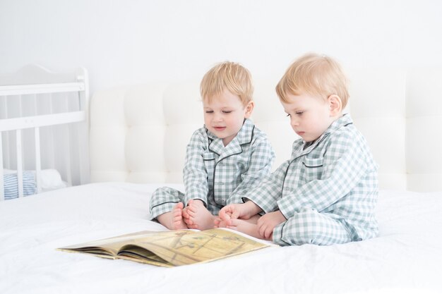 Twee peuterbaby tweelingjongens in pyjama's lezen boek zittend op wit beddengoed op bed