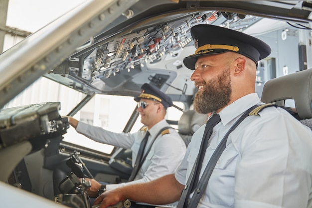 Foto twee positieve aantrekkelijke piloten die glimlachen in de cockpit van het vliegtuig terwijl ze zitten met hun veiligheidsgordels vastgemaakt