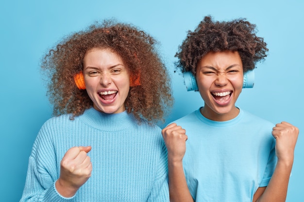 Foto twee positieve vrouwen van gemengd ras balden hun vuisten verheugen zich uitstekend nieuws voelt alsof winnaars genieten van het luisteren naar muziek via draadloze koptelefoons tijd samen doorbrengen, terloops gekleed geïsoleerd over blauwe muur