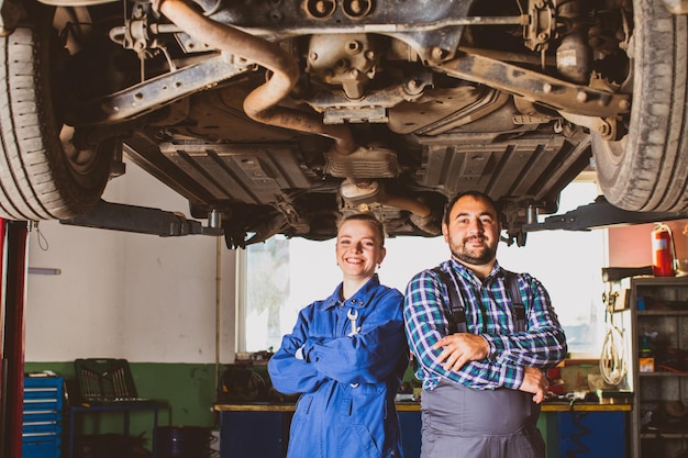 Twee professionele monteurs, een autoservicemedewerker, knappe man en jonge blonde vrouwen staan onder de hydraulische oprit met auto glimlachend naar de camera kijkend