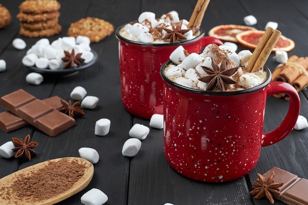 Twee rode kopjes warme chocolademelk met marshmallow, anijs en kaneel bestrooid met cacaopoeder