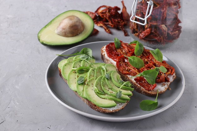 Twee sandwiches met avocado, gedroogde tomaten, rucola en erwten microgroen op grijze plaat op grijze betonnen ondergrond, close-up, ruimte voor tekst