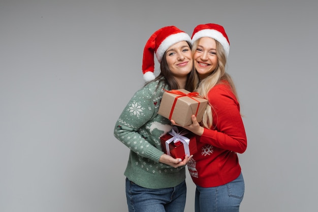 Twee santameisjes in rode hoeden met geschenken die nieuwjaar vieren en kerstmis poseren op een grijze achtergrond met kopieerruimte