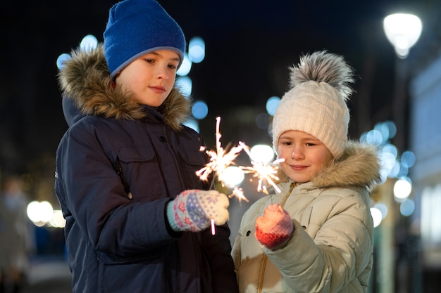 Twee schattige jonge kinderen, jongen en meisje in warme winter kleding met brandende sparkler vuurwerk op donkere nacht buiten. Nieuwjaar en kerstviering concept.