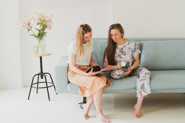 Twee schattige jonge vrouwen die thuis op de bank kletsen, koffie drinken en naar een modetijdschrift kijken