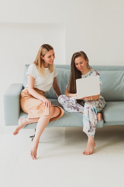 Twee schattige jonge vrouwen die thuis op de bank kletsen, koffie drinken en op een laptop werken