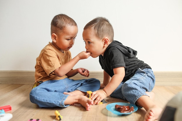 Twee schattige kleine kinderen eten samen koekjes terwijl ze op de vloer zitten Grappige Aziatische peutervoeding