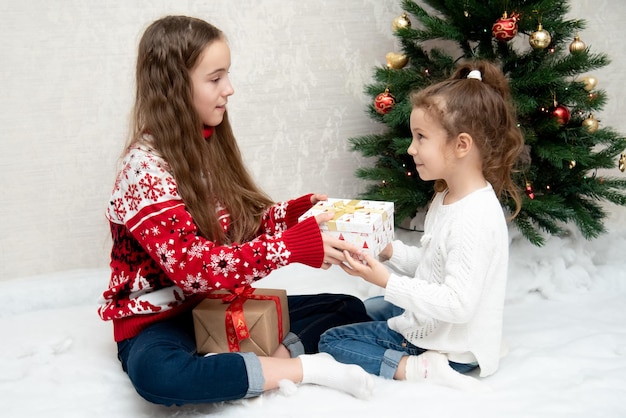 Twee schattige lachende meisjes zitten naast de kerstboom en geven elkaar nieuwjaarskerst cadeaus