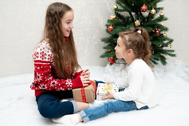 Twee schattige lachende meisjes zitten naast de kerstboom en geven elkaar nieuwjaarskerst cadeaus