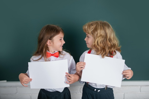 Twee schoolkinderen die een witte papieren blanco poster met kopieerruimte houden schoolmeisje en schooljongen in de klas