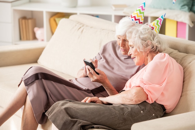 Twee senior femaled in verjaardag caps zittend op de Bank terwijl nieuwsgierige dingen in smartphone bespreken