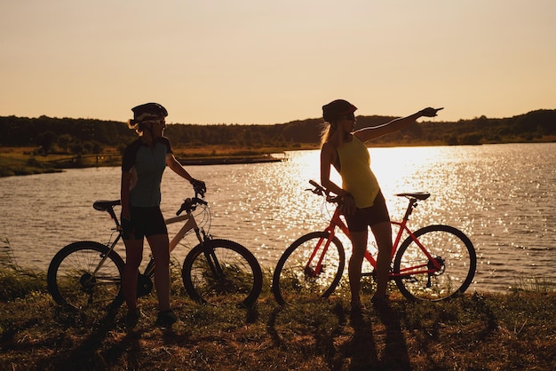 Twee silhouetten van vrouwen met een fiets die aan het meer staan en met elkaar praten bij de zonsondergang