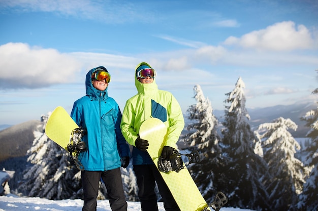 Twee snowboarders poseren in het skigebied Riders-vrienden die hun snowboards door het bos dragen voor backcountry freeride en een reflecterende bril dragen, kleurrijke mode-outfit Copyspace-gebied