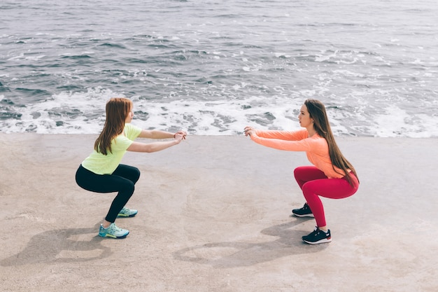 Twee sportmeisjes die squats op het strand doen.