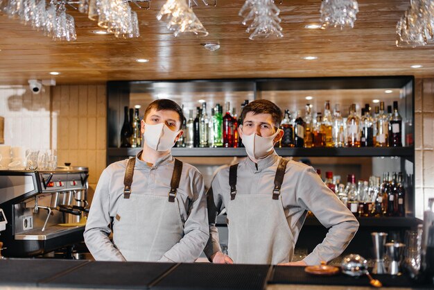 Twee stijlvolle barmannen in maskers en uniformen tijdens de pandemie staan achter de bar. het werk van restaurants en cafés tijdens de pandemie.