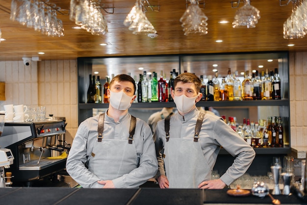 Twee stijlvolle barmannen in maskers en uniformen tijdens de pandemie staan achter de bar. Het werk van restaurants en cafés tijdens de pandemie.