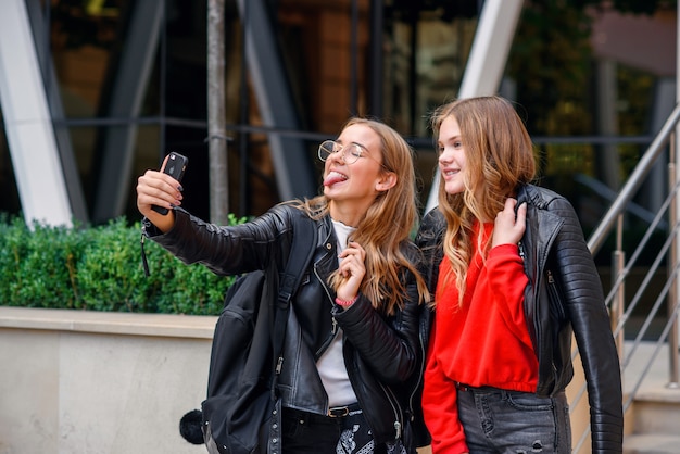 Twee stijlvolle gelukkig tienermeisjes met smartphone selfie maken in de buurt van stijlvol modern gebouw op straat.