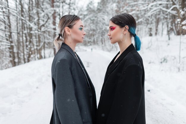 Twee stijlvolle mooie jonge vrouwenmodellen met een heldere uitstraling in vintage modieuze blazers staan tegenover elkaar in de winternatuur met sneeuw