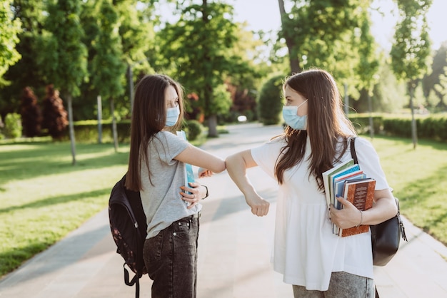 Twee studentmeisjes met beschermende medische maskers begroeten hun ellebogen wanneer ze elkaar ontmoeten op de campus. Onderwijs op afstand. Zachte selectieve focus.