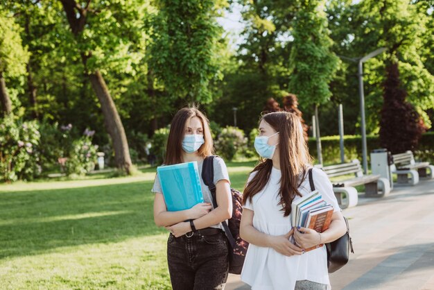 Twee studentmeisjes met beschermende medische maskers lopen en praten op de campus. Onderwijs op afstand. Zachte selectieve focus.