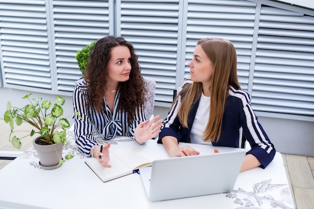 Twee succesvolle jonge zakelijke vrouwelijke managers zitten aan een tafel met een laptop en een notebook en werken aan een nieuw ontwikkelingsproject, vrouwelijke studenten schrijven een rapport over het werk van hun computer