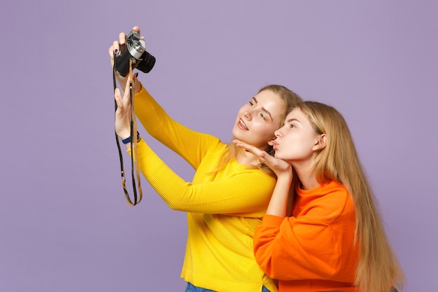 Twee tedere blonde tweelingzusters meisjes doen selfie geschoten op retro vintage fotocamera, blazen luchtkus geïsoleerd op violet blauwe muur. Mensen familie levensstijl concept.
