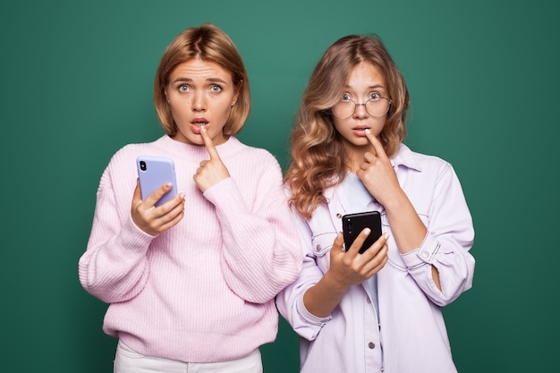 Twee teleurgestelde blonde vrouwen die op een groene muur chatten, raken haar lippen met vinger aan en kijken direct verdrietig