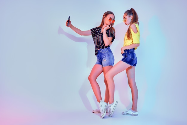Twee tienermeisjes in zomerkleren permanent in studio en selfie maken op witte achtergrond