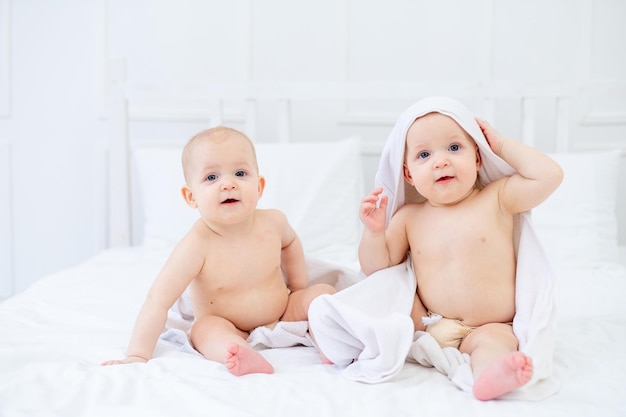 Twee tweelingbaby's in een witte handdoek na het baden in een badkuip op een helder bed thuis lachend met het concept van hygiëne en wassen