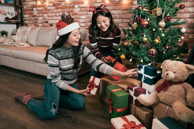 twee van jonge vrouwelijke vrienden naast de kerstboom ingepakte cadeautjes onder zetten en plezier hebben. gelukkige meisjes in santa-rendierhoeden die huisgeschenkdozen versieren en teddybeer voorbereid op kerstvakantie