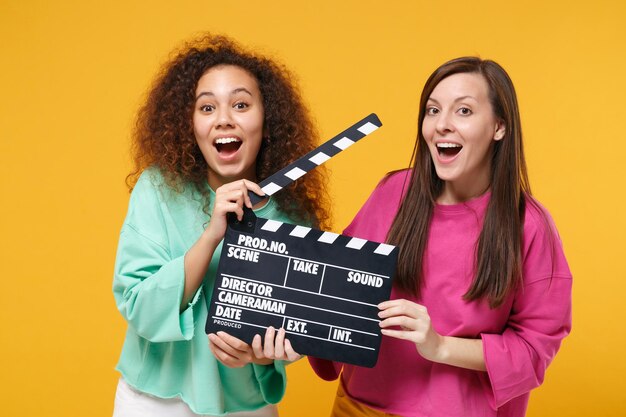 Twee verbaasde vrouwen vriend Europese Afrikaanse Amerikaan in roze groene kleding poseren geïsoleerd op gele achtergrond. Mensen levensstijl concept. Bespotten kopie ruimte. Houd klassieke filmklapper vast.